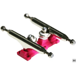 May include: A pair of pink fingerboard trucks with silver axles and wheels. The trucks are designed for use with fingerboards, which are miniature skateboards.