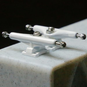 May include: Two white miniature skateboard trucks are displayed on a speckled surface. The trucks have silver hardware and are designed for fingerboards. The trucks are painted white and have a clean, modern aesthetic.