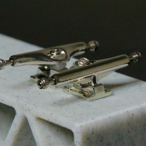 May include: A pair of silver fingerboard trucks. The trucks are mounted on a white surface with black flecks.