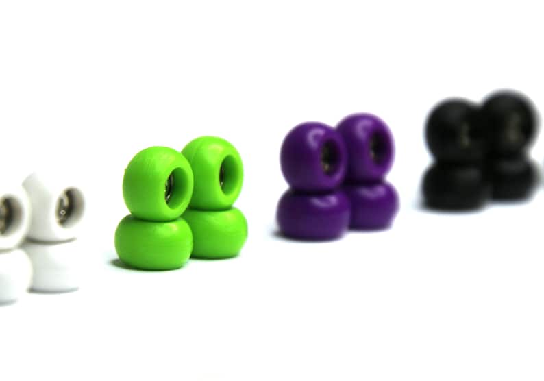 May include: Set of four pairs of fingerboard wheels in white, green, purple, and black. Each pair is stacked on top of each other.