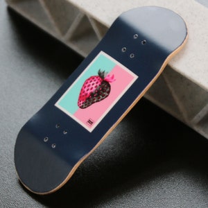 May include: A dark blue fingerboard with a graphic of a pink strawberry on a light blue and pink background. The fingerboard has a wood deck and black wheels.
