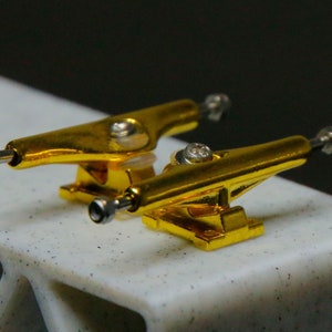 May include: Two gold-coloured fingerboard trucks with silver axles and wheels. The trucks are mounted on a white surface.