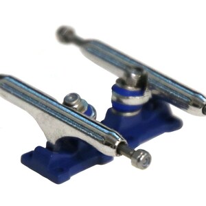 May include: A pair of silver fingerboard trucks with blue baseplates and silver hardware. The trucks are designed for fingerboards and are made of metal.