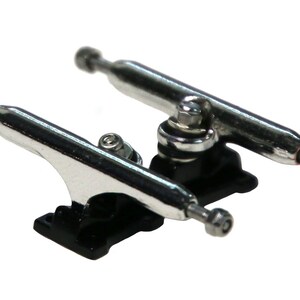 May include: A pair of silver fingerboard trucks with black baseplates. The trucks are designed for fingerboards and have a classic skateboard truck design.