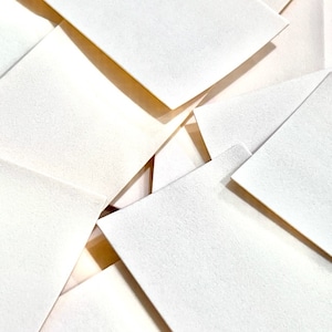 Op de afbeelding: Een stapel witte enveloppen, sommige zijn gevouwen en sommige zijn plat, met een gestructureerd oppervlak.