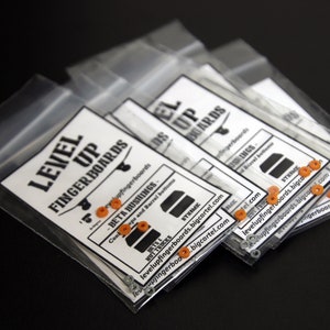 May include: A close-up of several small plastic bags containing orange and silver fingerboard parts. The bags are labeled "Level Up Fingerboards" and include the website address "levelupfingerboards.bigcartel.com".