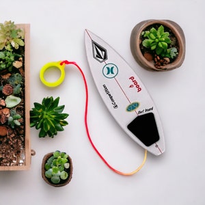 Puede incluir: Tabla de surf blanca con gráficos negros y turquesas, una correa roja y un anillo amarillo. La tabla tiene las palabras "board" y "Surf board" impresas. Rodeada de pequeñas suculentas en macetas.