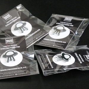 Puede incluir: Cuatro bolsas de plástico transparentes que contienen cada una un pin de coleccionista de edición limitada en blanco y negro con un OVNI plateado de tres patas. Las bolsas están etiquetadas con "Pin de coleccionista de edición limitada" y "www.fingerspace.co".