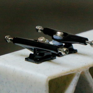 May include: Two black fingerboard trucks with silver hardware. The trucks are mounted on a white surface.