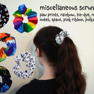 Puede incluir: Una colección de scrunchies coloridos con varios patrones, incluyendo huellas de patas, arcoíris, tie-dye, notas musicales, espacio y lunares.