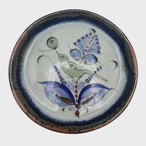 May include: Round ceramic plate with a hand-painted design. The plate features a light blue and white background with a dark blue and brown rim. A green bird is perched on a blue floral design, with a butterfly above it. The plate is a decorative item.