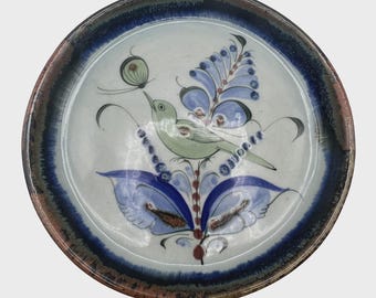 Könnte beinhalten: Runder Keramikteller mit handgemaltem Design. Der Teller hat einen hellblauen und weißen Hintergrund mit einem dunkelblauen und braunen Rand. Ein grüner Vogel sitzt auf einem blauen Blumendesign, mit einem Schmetterling darüber. Der Teller ist ein dekorativer Artikel.