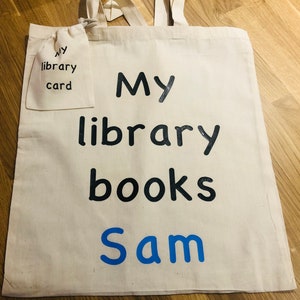 May include: Cream-colored canvas tote bag with the words "My library books Sam" in black and blue. A smaller matching pouch is attached, labeled "My library card". The bag has two handles.