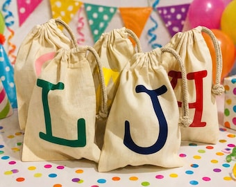 May include: Beige drawstring bags with large, colorful letters. The bags have a natural, textured appearance and are arranged in a group. Party hats, balloons, and a wrapped gift box are in the background, suggesting a celebration or event.
