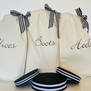 Puede incluir: Tres bolsas de lona blancas con lazos de cinta rayados en blanco y negro. Las bolsas están etiquetadas como "Shoes", "Boots" y "Heels". También se incluye una cinta rayada en blanco y negro.