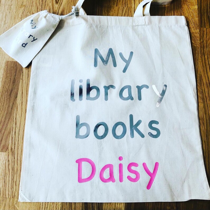 May include: A cream-colored canvas tote bag with the words "My library books Daisy" printed in blue and pink. A matching small pouch with the words "My library" is attached to the bag.