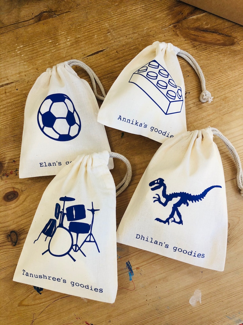May include: Four white drawstring bags with blue designs and names. The bags are on a wooden surface. The designs include a soccer ball, a Lego brick, a dinosaur, and a drum set. The bags are labeled with the names "Elan's goodie", "Annika's goodie", "Dhilans goodies", and "Tanushree's goodies".