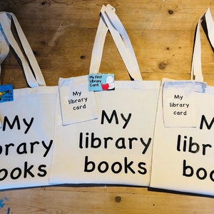 May include: Three off-white canvas tote bags with long handles. Each bag has the words "My library books" printed in black. Each bag has a small pocket with "My library card" printed on it. One bag has a small card that says "My First Library Card".