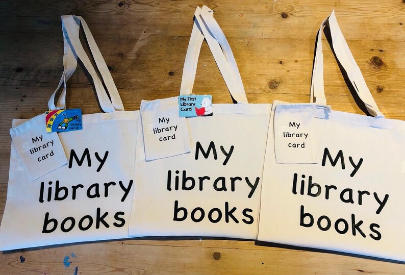 May include: Three off-white canvas tote bags with long handles. Each bag has the words "My library books" printed in black. Each bag has a small pocket with "My library card" printed on it. One bag has a small card that says "My First Library Card".