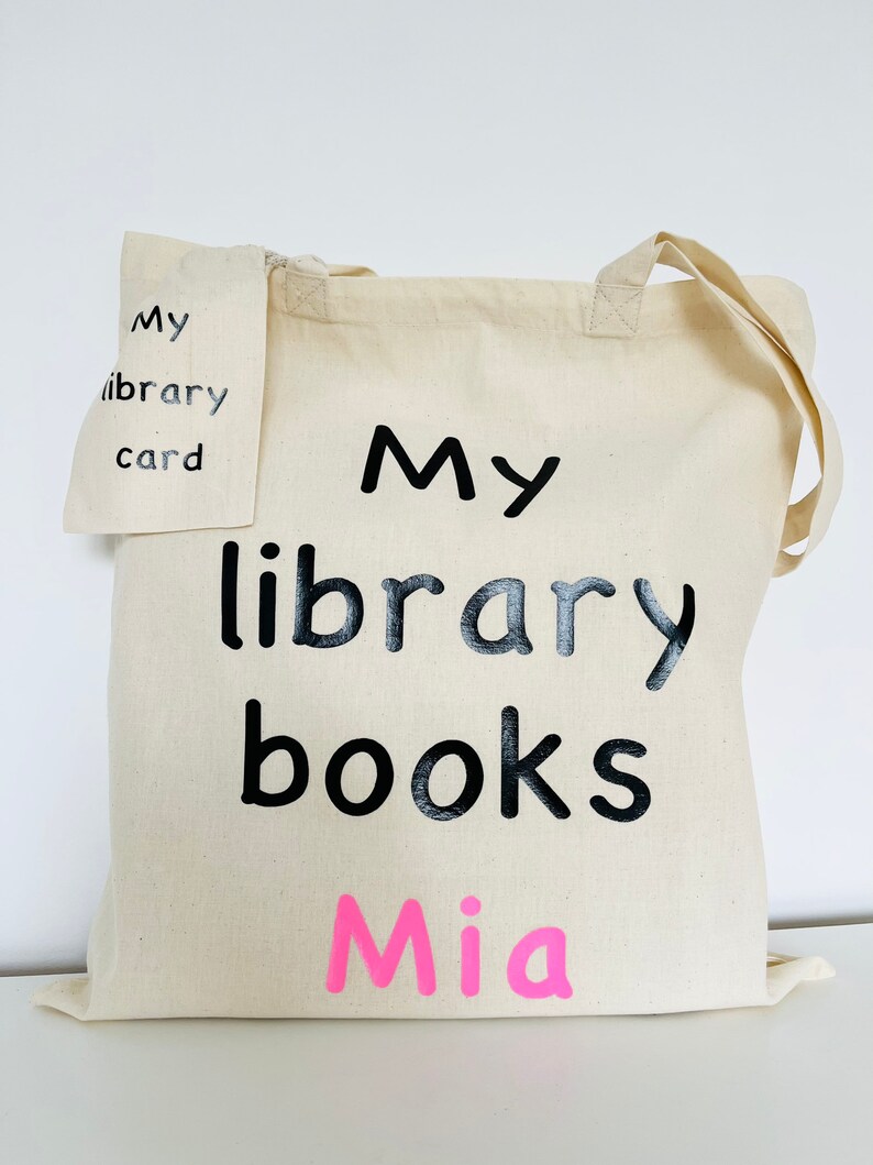May include: A natural canvas tote bag with black text that reads "My library books Mia." A smaller matching pouch reads "My library card." The bag has two handles and is designed for carrying books.