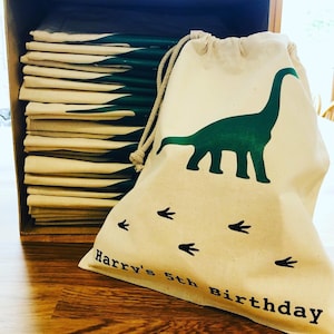 May include: A stack of 15 natural canvas drawstring bags with a green dinosaur and footprints design. The bag in the front has the text "Harry's 5th Birthday" printed on it.