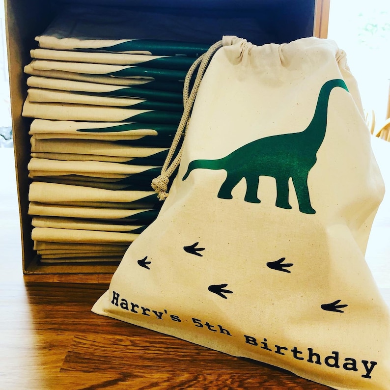 May include: A stack of 15 natural canvas drawstring bags with a green dinosaur and footprints design. The bag in the front has the text "Harry's 5th Birthday" printed on it.