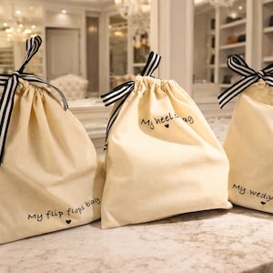 Op de afbeelding: Drie beige trekkoordtassen met zwart-wit gestreepte linten. Elke tas heeft handgeschreven tekst: "My flip flops bag", "My heels bag" en "My wedges bag". De tassen staan op een marmeren oppervlak.