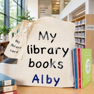 May include: A beige canvas tote bag with the words "My library books Alby" printed on it in black and blue. A small matching pouch reads "My library card." Several colorful books are next to the bag, with the word "Fiction" visible on the spines.