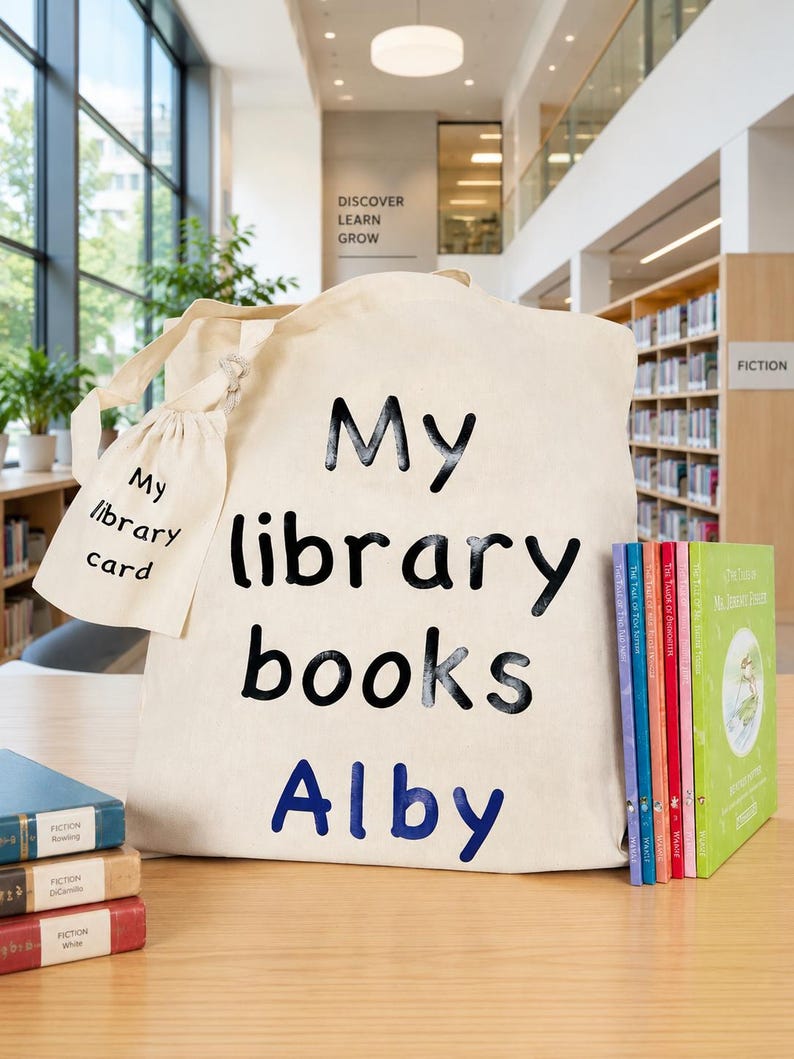 May include: A beige canvas tote bag with the words "My library books Alby" printed on it in black and blue. A small matching pouch reads "My library card." Several colorful books are next to the bag, with the word "Fiction" visible on the spines.