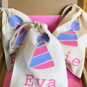 May include: Three white cotton drawstring bags with pink and blue party hats and names printed on them. The bags are in a cardboard box with pink tissue paper.