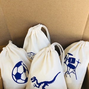 May include: Four white drawstring bags with blue designs. The designs include a soccer ball, a dinosaur skeleton, a drum set, and a set of building blocks. The bags are in a cardboard box.