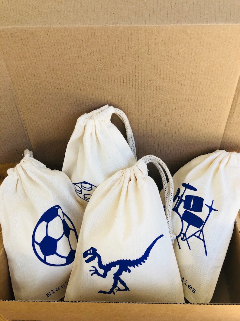 May include: Four white drawstring bags with blue designs. The designs include a soccer ball, a dinosaur skeleton, a drum set, and a set of building blocks. The bags are in a cardboard box.
