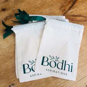 May include: Three white cotton drawstring bags with green ribbon ties. The bags have a green logo that says "Bodhi Aspirations" with a small leafy branch design.