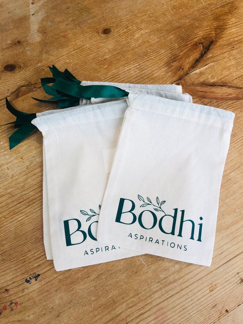 May include: Three white cotton drawstring bags with green ribbon ties. The bags have a green logo that says "Bodhi Aspirations" with a small leafy branch design.