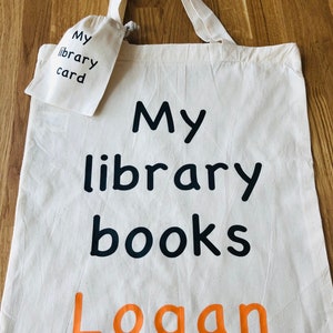May include: Off-white canvas tote bag with black text reading "My library books" and the name "Logan" in orange. A small matching pouch with "My library card" is attached. The bag has long straps.