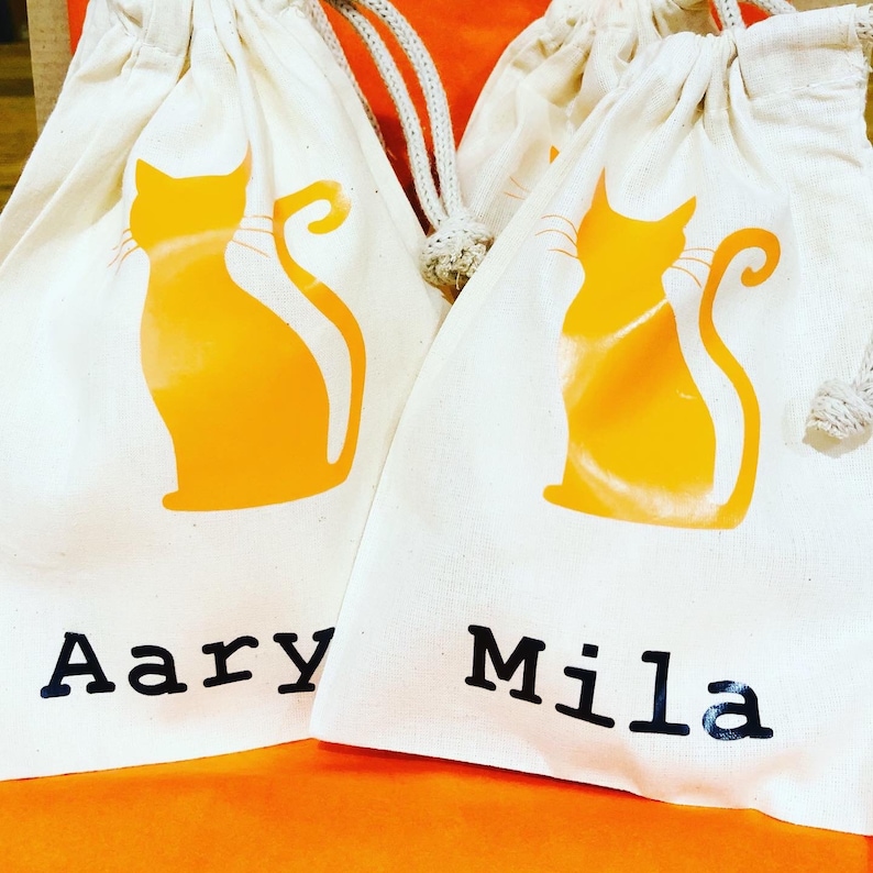 May include: Two white cotton drawstring bags with orange cat silhouettes and the names "Aary" and "Mila" printed in black.