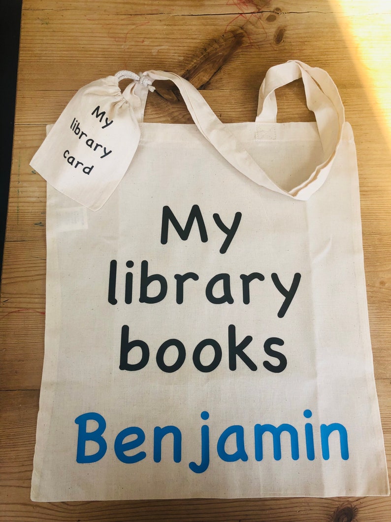 May include: A cream-colored tote bag with the text "My library books Benjamin" in black and blue. A small matching pouch with "My library card" is attached. The bag has two handles and is made of a sturdy fabric.