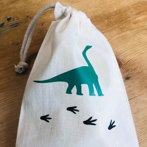 May include: A white cotton drawstring bag with a green dinosaur silhouette and black dinosaur footprints. The bag has the text "August's 4th Birthday" printed on the bottom.
