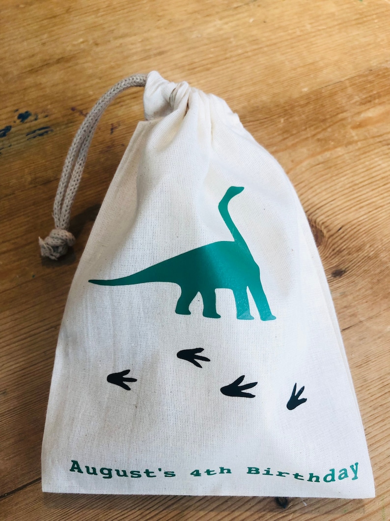 May include: A white cotton drawstring bag with a green dinosaur silhouette and black dinosaur footprints. The bag has the text "August's 4th Birthday" printed on the bottom.