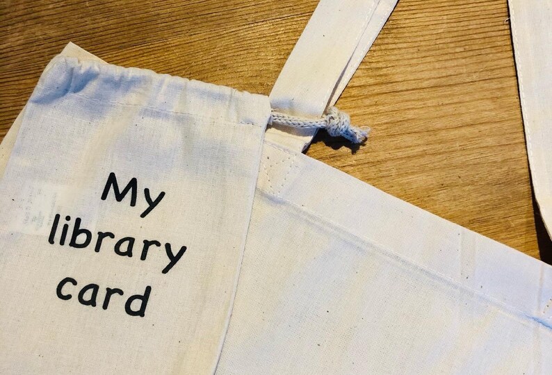 May include: A cream-colored canvas bag with a drawstring closure. The words "My library card" are printed in black, bold letters on the front of the bag. The bag has a simple, minimalist design.