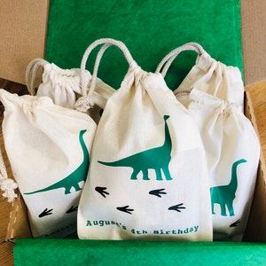May include: A set of four white cotton drawstring bags with green dinosaur designs and the text "August's 4th Birthday". The bags are in a cardboard box lined with green tissue paper.