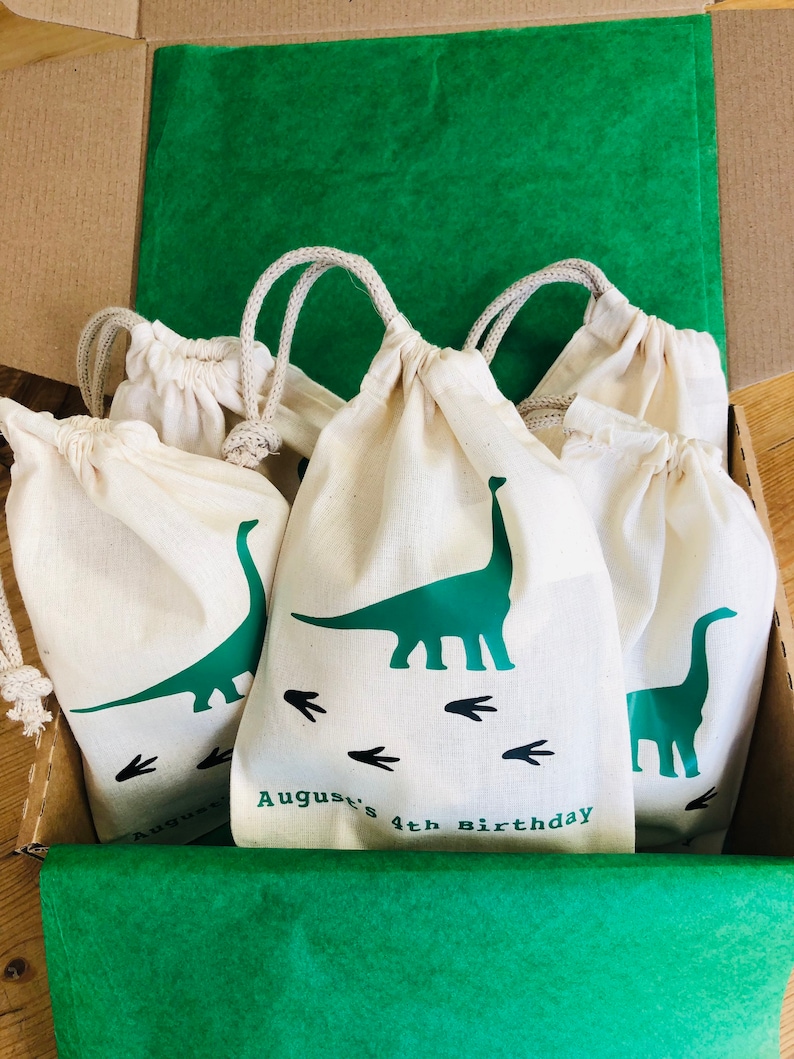 May include: A set of four white cotton drawstring bags with green dinosaur designs and the text "August's 4th Birthday". The bags are in a cardboard box lined with green tissue paper.