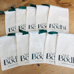 May include: Ten white cotton drawstring bags with green ribbon ties. Each bag has a green logo that says "Bodhi Aspirations" with a small leafy branch design.