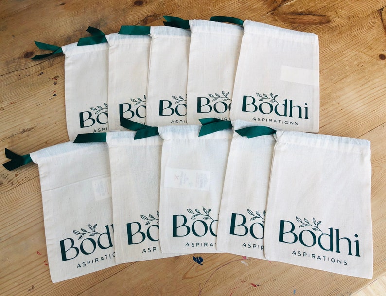 May include: Ten white cotton drawstring bags with green ribbon ties. Each bag has a green logo that says "Bodhi Aspirations" with a small leafy branch design.