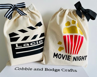 May include: Two white canvas drawstring bags with black designs. One bag has a black and white clapperboard with the name "Lacie" on it. The other bag has a red and yellow popcorn bucket with the text "Movie Night".