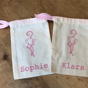 May include: Two white cotton drawstring bags with pink ribbon ties. Each bag has a pink silhouette of a cat and a name printed on the front. The names are "Sophie" and "Klara".