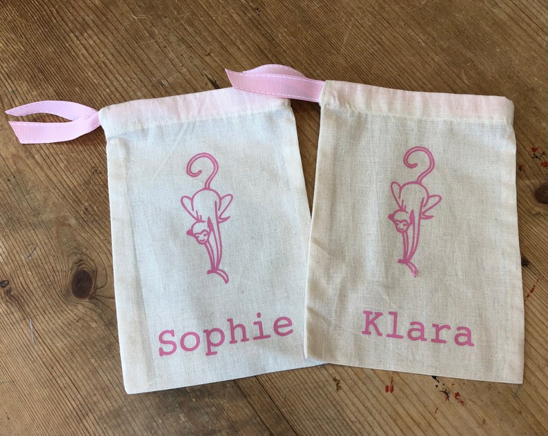 May include: Two white cotton drawstring bags with pink ribbon ties. Each bag has a pink silhouette of a cat and a name printed on the front. The names are "Sophie" and "Klara".