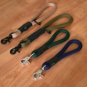 Puede incluir: Cuatro correas para perros de diferentes colores con asas negras y clips plateados. Las correas están hechas de cuerda y están dispuestas en fila sobre una superficie de madera.