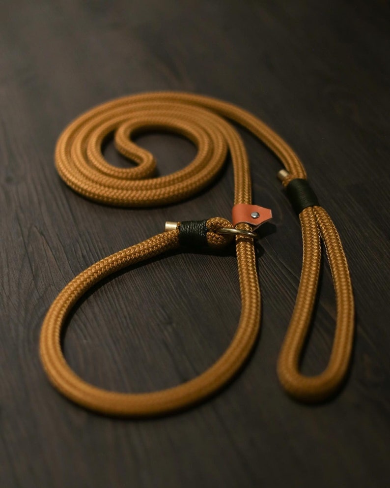 May include: A tan rope dog leash with a loop handle and a loop for the dog's neck. The leash has a leather accent and metal hardware. The rope is thick and appears durable, suitable for medium to large dogs.