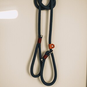 May include: A navy blue dog leash with a loop handle and a metal clasp. The leash is made of thick, braided rope and features brown leather accents. The leash is hanging on a white wall.